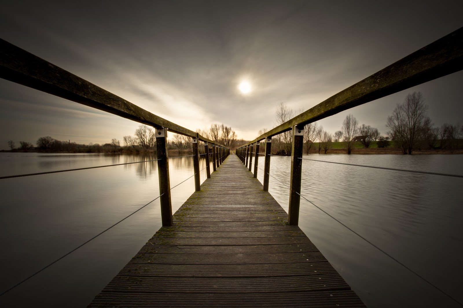 A wooden pier stretches out into calm water under a cloudy sky with the sun faintly visible.